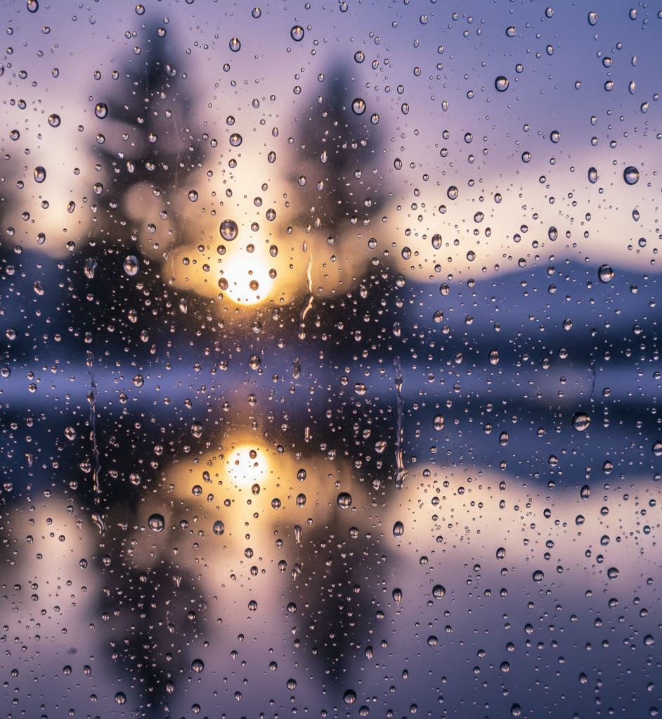 Gotas de chuva escorrendo em um vidro com o reflexo de um sol poente sobre um lago e árvores ao fundo, simbolizando esperança, limpeza da alma e o propósito das provações.